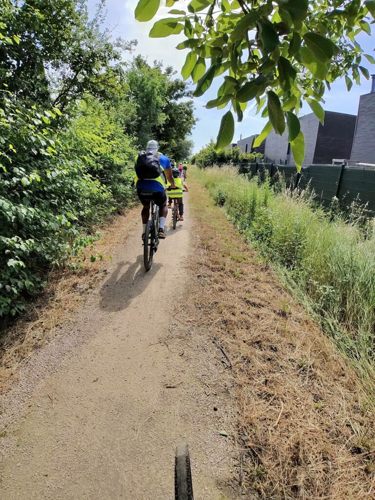 Groupe de cyclistes en gilets jaunes sur un chemin de terre bordé de verdure à Pusignan, pendant la sortie Puz à Vélo